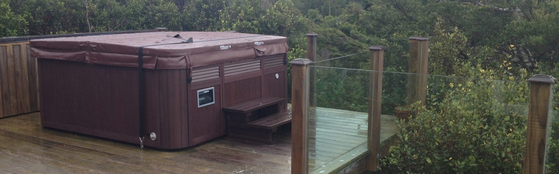 Spa with wind straps on a wet timber deck beside a glass fence